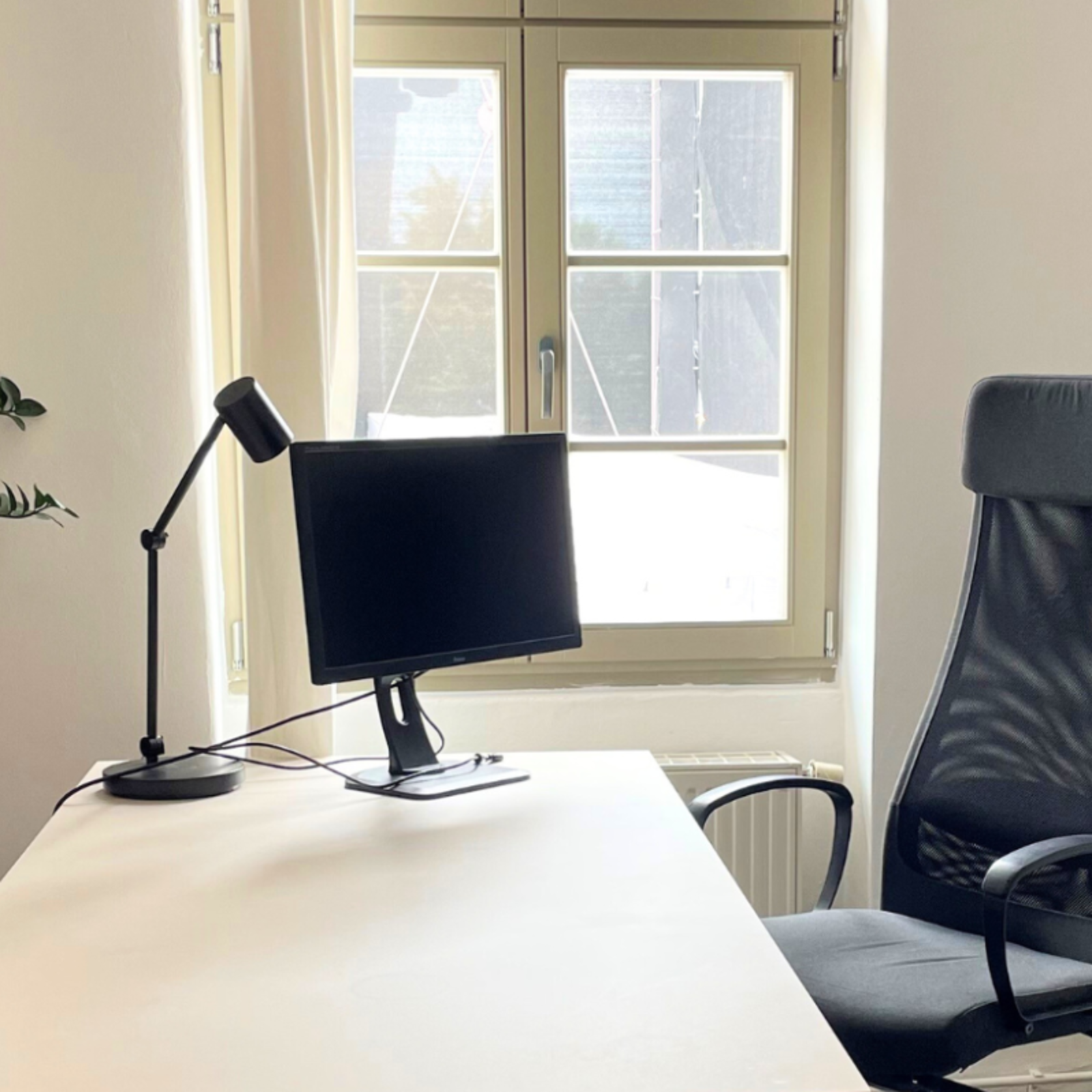 Moderner Arbeitsplatz mit weißem Schreibtisch, schwarzem Bürostuhl, Monitor, Schreibtischlampe, zwei Pflanzen und Fenster im Hintergrund
