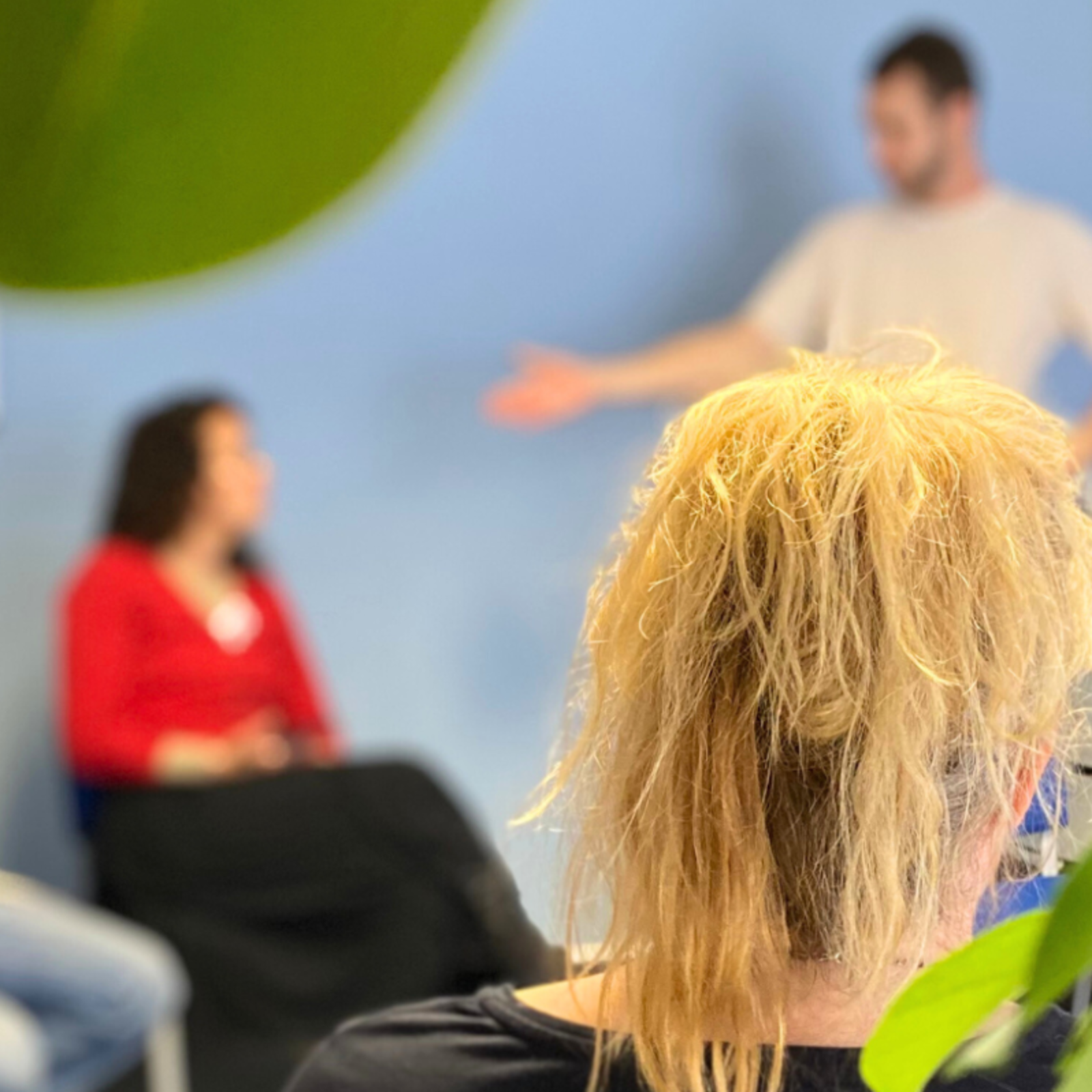 Person mit blonden, zusammengebundenen Haaren sitzt in einem Stuhl, im Hintergrund drei sitzende Personen und eine stehende Person in einem Raum mit blauen Wänden.